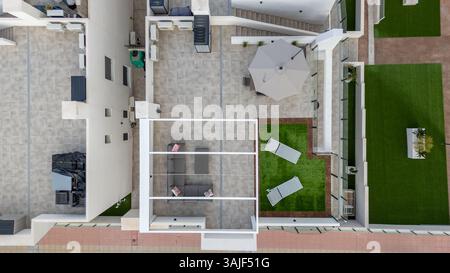 Drohnenaufnahme eines Dachs mit Einzelsitzen, Tisch, Sonnenschirm und Kunstrasenbereich mit Sonnenliegen in einem modernen Apartmentkomplex. Stockfoto