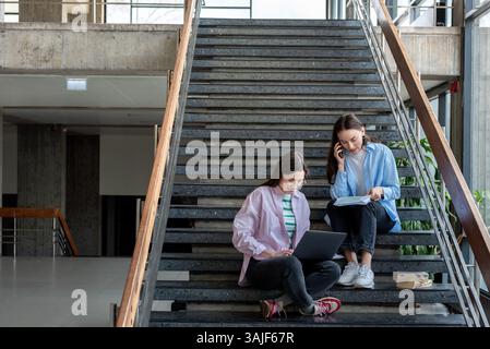 Zwei Studentinnen arbeiten nebeneinander, während sie auf der Treppe in der Lobby der Universität sitzen. Stockfoto