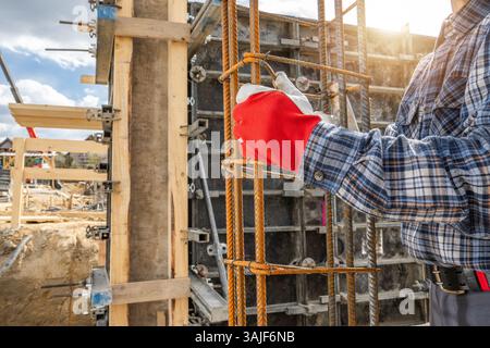 Ein Bauarbeiter, der Handschuhe und ein kariertes Hemd trägt, greift Metallbewehrung auf einer Baustelle. Die Sonne scheint hell im Hintergrund, Highlighti Stockfoto