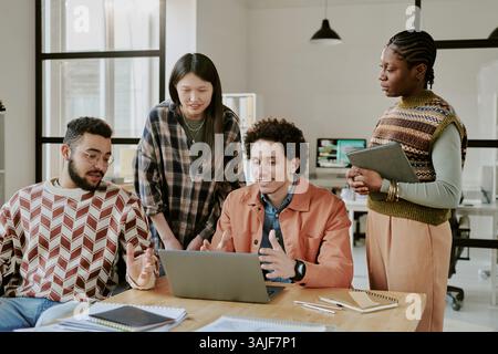 Begeisterte hochqualifizierte Programmierer, die sich ein Notebook ansehen und während des Arbeitstages in einem geräumigen Büro lebhafte Diskussionen führen Stockfoto