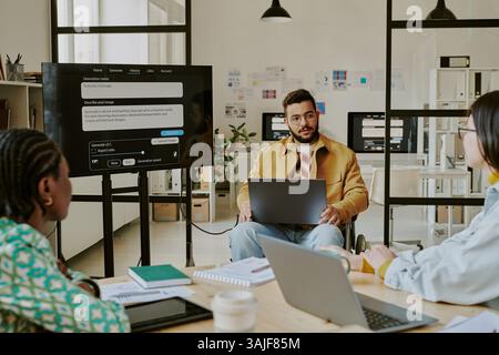 Selbstbewusster Teamleiter mit Behinderung hält eine Präsentation vor IT-Spezialisten, die mit Bürobedarf und digitalen Geräten am Holztisch sitzen Stockfoto