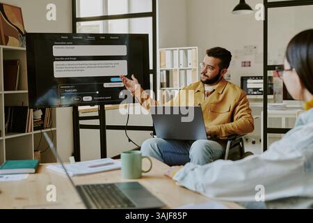 Nicht erkennbare IT-Fachfrau sitzt am Holztisch und hört dem Teamleiter aufmerksam zu. Professionelle Teamleiterin mit Behinderung hält Präsentation im Büro Stockfoto