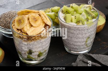 Zwei Gläser Chiasamenpudding, eines mit Kiwi-Würfel und das andere mit Müsli und Bananenchips, befinden sich auf einer dunklen Oberfläche mit Zutaten Stockfoto