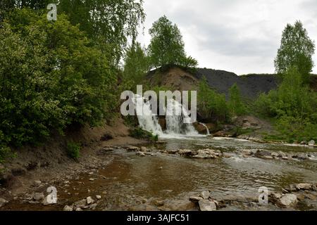 Ein kleines Rückwasser in der Nähe eines hohen Wasserfalls, der mit einem schnellen Bach in die Felsen fließt, umgeben von einem Birkenwald an einem bewölkten Sommertag. Bel Stockfoto