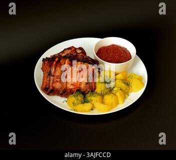 Gebackene Schweinerippchen mit Tomatenwürzsauce und jungen gekochten Kartoffeln auf einer großen weißen Keramikplatte auf schwarzem Hintergrund. Nahaufnahme. Stockfoto