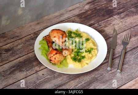 Zwei gebackene Hähnchenschenkel auf einem Salatblatt und Kartoffelpüree mit grünen Zwiebeln auf einem Holztisch, Besteck in der Nähe. Nahaufnahme. Stockfoto