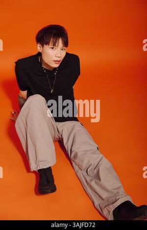 Selbstbewusster junger asiatischer Mann in trendigem Outfit sitzt auf dem Boden vor einem leuchtend orangen Hintergrund. Stockfoto