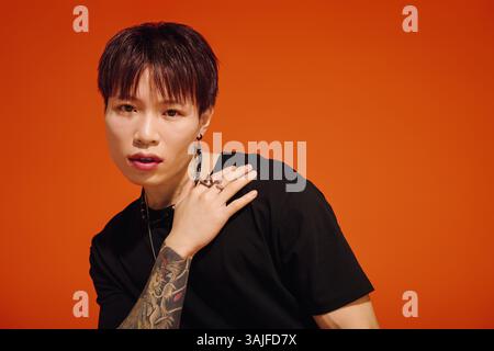 Ein stilvoller junger asiatischer Mann zeigt seine Mode und Tattoos vor einem auffälligen orangen Hintergrund. Stockfoto
