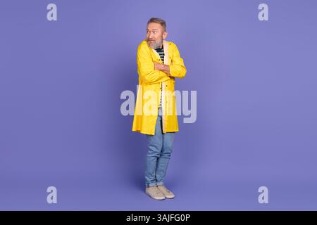 Älterer Mann mit stilvollem Bart in gelbem Regenmantel und lässigem Outfit auf violettem Hintergrund, das Lifestyle und Mode zeigt Stockfoto
