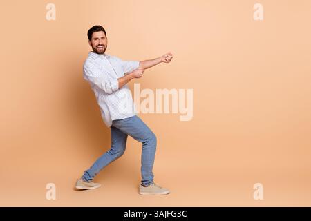 Ein lächelnder junger Mann posiert in einem stilvollen, legeren Outfit vor beigefarbenem Hintergrund mit einer verspielten Geste, an einem imaginären Seil zu ziehen. Stockfoto