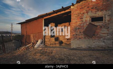Rustikales Scheuneninnere mit Heuballen, die von goldenem Sonnenuntergang beleuchtet werden, und der Silhouette der Stadt in der Ferne vor warmem Abendlicht Stockfoto