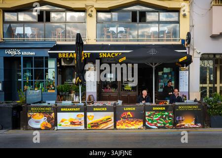 St. Julian’s, Malta- 09.04.2025: Vormittag in einem Straßencafé auf Malta. Die Menschen genießen Kaffee auf der Außenterrasse, umgeben von Essensbanner. Stockfoto