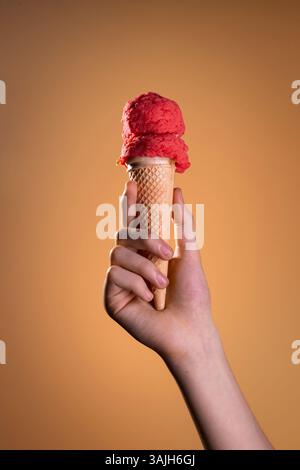 Erdbeer-Eiskegel von Hand auf orangem Hintergrund gehalten. Sommerdessert, lebhaftes und erfrischendes Konzept mit rotem Eislöffel aus nächster Nähe. Stockfoto
