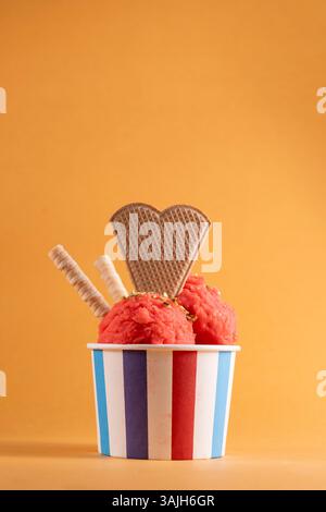 Erdbeereis in farbenfroher Tasse mit herzförmiger Waffel und gerollten Waffelstäbchen auf orangefarbenem Hintergrund. Frisches und lebhaftes Sommerdessert. Stockfoto