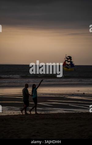 Bali, Indonesien, 11. November 2024: Eine ruhige Strandszene mit einem Paar, das während des Sonnenuntergangs einen Spaziergang macht, während es einen lebhaften Drachen fliegt. Stockfoto