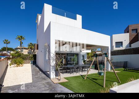 Hinter der Fassade einer stilvollen modernen Villa mit Kunstrasen, Kinderschaukel und überdachter Terrasse mit großem Esstisch und Stühlen. Stockfoto