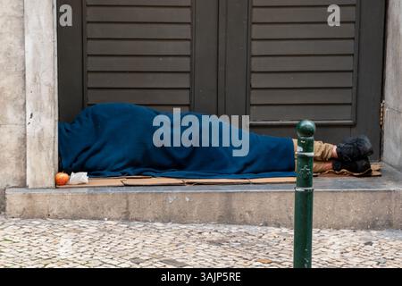 Mann ohne Haus schläft am ruhigen Strand, liegt auf Sand mit abgenutzter Decke, Meer im Hintergrund, friedliche Küstenszene Stockfoto