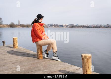 Eine junge Frau in einer orangefarbenen Jacke sitzt auf einem hölzernen Pier und blickt an einem sonnigen Tag auf einen ruhigen See und eine ferne Stadtlandschaft. Stockfoto