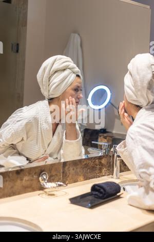 Eine Frau im Bademantel wendet Hautpflege auf ihr Gesicht an, während sie in einen gut beleuchteten Spiegel blickt. Ihr Haar ist in ein Handtuch gewickelt, und im Badezimmer ist Ele Stockfoto