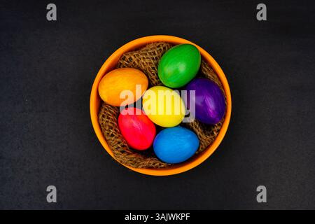 Bunte Ostereier in Orange Schüssel auf schwarzem Hintergrund. Bunte festliche Eier in einer strukturierten orangen Schüssel mit Garn, gestylt auf einem dunklen B Stockfoto