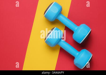 Plastikhanteln auf rot-gelbem Papierhintergrund. Minimalistisches Fitnesskonzept. Draufsicht Stockfoto