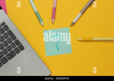Laptop mit Stiften und Notizpapier mit Fragezeichen auf gelbem Hintergrund. Draufsicht Stockfoto