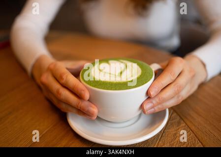 Frauenhände, die eine Tasse Matcha Latte mit herzförmigem Design in cremigem Schaum auf einem Holztisch liegen, während sie ein warmes, wohltuendes Getränk genießen Stockfoto