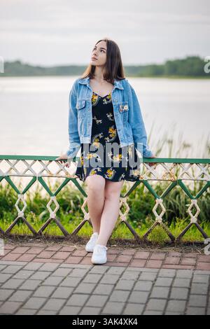 Junge Frau, die sich auf das Geländer am See lehnt und eine ruhige Wasserlandschaft mit entspannter Haltung betrachtet Stockfoto