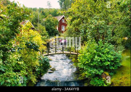 Bach mit Einer kleinen Brücke vor Einem Roten Holzhaus in Schweden. Bäume Auf Der Bank . Skandinavische Natur Stockfoto