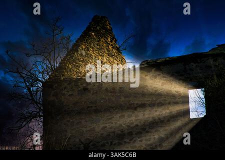 Ein altes Steingebäude steht teilweise beleuchtet von Lichtstrahlen, die durch eine Tür brechen und sich in der Dämmerung gegen dunkle, wirbelnde Wolken setzen. Stockfoto