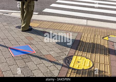 Lange Schatten und große Cargo Hosen sind der Mittelpunkt der urbanen Szene mit jungen Frauen, die auf taktilen Pflastersteinen auf Zebras gestreiftem Crosswalk in Japan stehen. Stockfoto