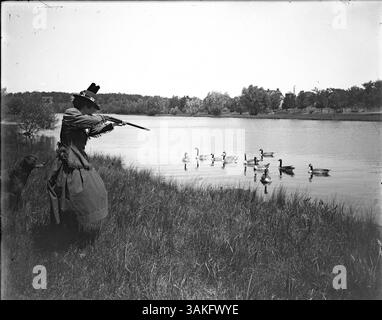 Nettie Shattuck, die erste Frau im Minneapolis Gun Club, sieht man Enten am Lake of the Isles schießen. Das Foto zeigt sie an der nördlichen Bucht des Sees mit einem Bauernhof auf der anderen Seite des Wassers. Ein Hund steht neben ihr, während er in diesem historischen Moment posiert. BR0970 Stockfoto