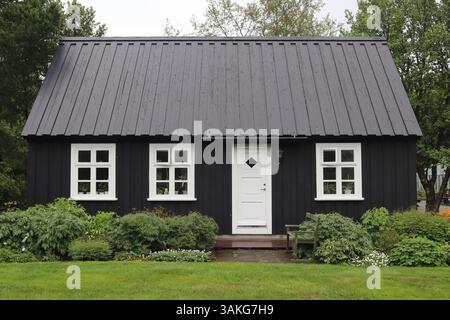 Seydisfjordur, Island - 13. Juni 2024: Schwarzes Holzhaus mit weißen Fenstern und Tür, eingebettet zwischen grünem Gras und Büschen in Seydisfjordur, Island Stockfoto
