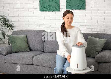 Junge Frau, die auf schwarzem Sofa sitzt und im Wohnzimmer modernen Luftbefeuchter anschaltet Stockfoto