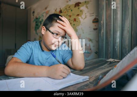 asiatischer Junge mit einer Brille, die frustriert den Kopf hält, Schulaufgabe am Heimschreibtisch in der Nähe der Welt, Konzept der Homeschooling, Bildungsdienstleistungen Stockfoto