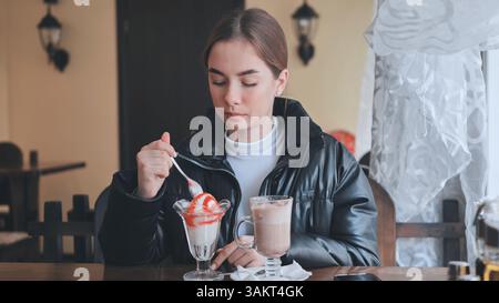 Eine junge Frau isst Eis und schlürft Latte, entspannt sich in einem gemütlichen Café mit süßem Genuss Stockfoto