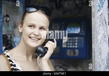 Attraktive Frau mit ihrer Sonnenbrille auf dem Kopf stehend mit einem öffentlichen Telefon mit einem Lächeln der Freude auf ihrem Gesicht Stockfoto