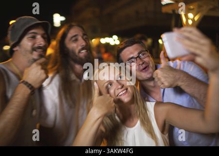 Eine Gruppe von glücklichen und aufgeregten jungen Leuten, die nachts im Freien Spaß haben. Konzentrieren Sie sich darauf, dass Mann und Frau die Daumen nach oben zeigen und ein Handy-Selfie machen Stockfoto