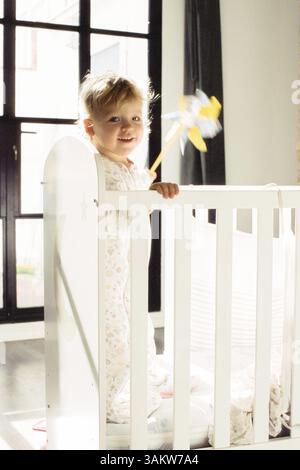 Startseite Porträt von glücklichen Baby Mädchen in Schlafbekleidung mit Spielzeug in der Krippe stehen, Blick in hellen Morgensonne Stockfoto