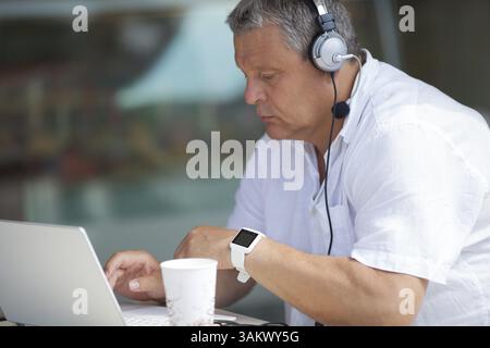 Älterer Mann in weißem T-Shirt und Kopfhörern, der am Laptop sitzt, während er im Café im Freien am Tisch sitzt Stockfoto