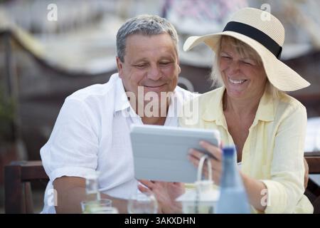 Lächelndes, glückliches, entspanntes Paar mittleren Alters, das im Urlaub zusammen ein Tablet benutzt, während sie an einem Tisch in einer Cafeteria sitzen und Erfrischungen genießen Stockfoto