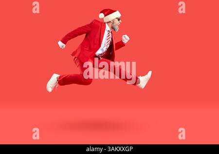 Alter Mann im Weihnachtsmann-Stil, Turnschuhe und Weihnachtsmütze, der in der Luft auf rotem Hintergrund schnell hochläuft. Stockfoto