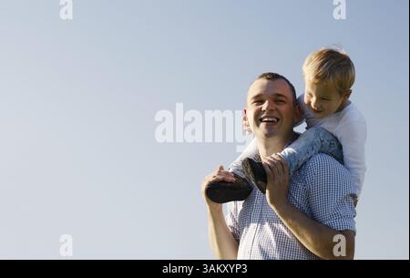 Junger Vater seinen Sohn eine piggy zurück fahren, da der Knabe sitzt auf seinen Schultern Lachen mit Genuss, Low Angle gegen einen blauen Himmel mit copysp Stockfoto