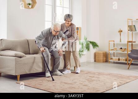Eine ältere Frau, die ihrem Mann hilft, mit Gehstock im Wohnzimmer zu Hause zu gehen. Stockfoto