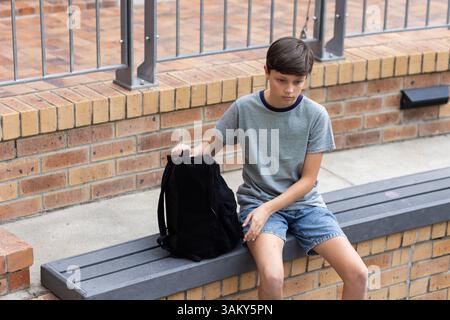Sitzt auf der Bank, der Junge hält den Rucksack und sieht nachdenklich in der Schule aus Stockfoto
