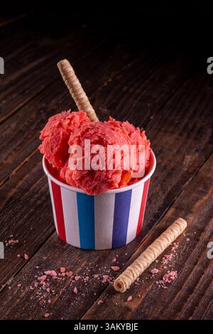 Zwei Messlöffel lebendiges Erdbeersorbet, serviert in gestreiftem Papierbecher über rustikaler Holzoberfläche, ideale Sommererfrischung Stockfoto