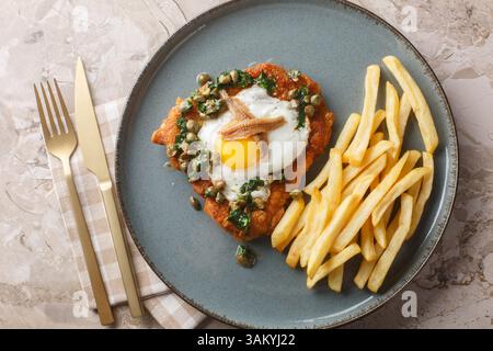 Köstliches paniertes Schnitzel mit Sardellen, Spiegelei und Kapern Petersiliensauce, serviert mit pommes Frites in Nahaufnahme auf einem Teller auf dem Tisch. Horiz Stockfoto