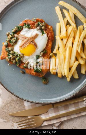Holsteinschnitzel mit Sardellenfilet, Spiegelei und Kapern-Petersiliensauce darauf serviert mit Pommes Frites in Nahaufnahme auf einem Teller auf einem Tisch. Vertikal oben Stockfoto