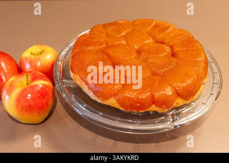 Blick von oben auf hausgemachte Tarte Tatin mit ordentlich angeordneten karamelisierten Apfelscheiben auf goldener Kruste, präsentiert mit ganzen Äpfeln auf neutralem Hintergrund Stockfoto