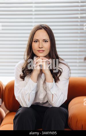Eine junge Frau in Bürokleidung sitzt anmutig und lehnt sich an ihre Hände. Sie strahlt Vertrauen, Professionalität und Erfolg in einem modernen Geschäftsumfeld aus Stockfoto